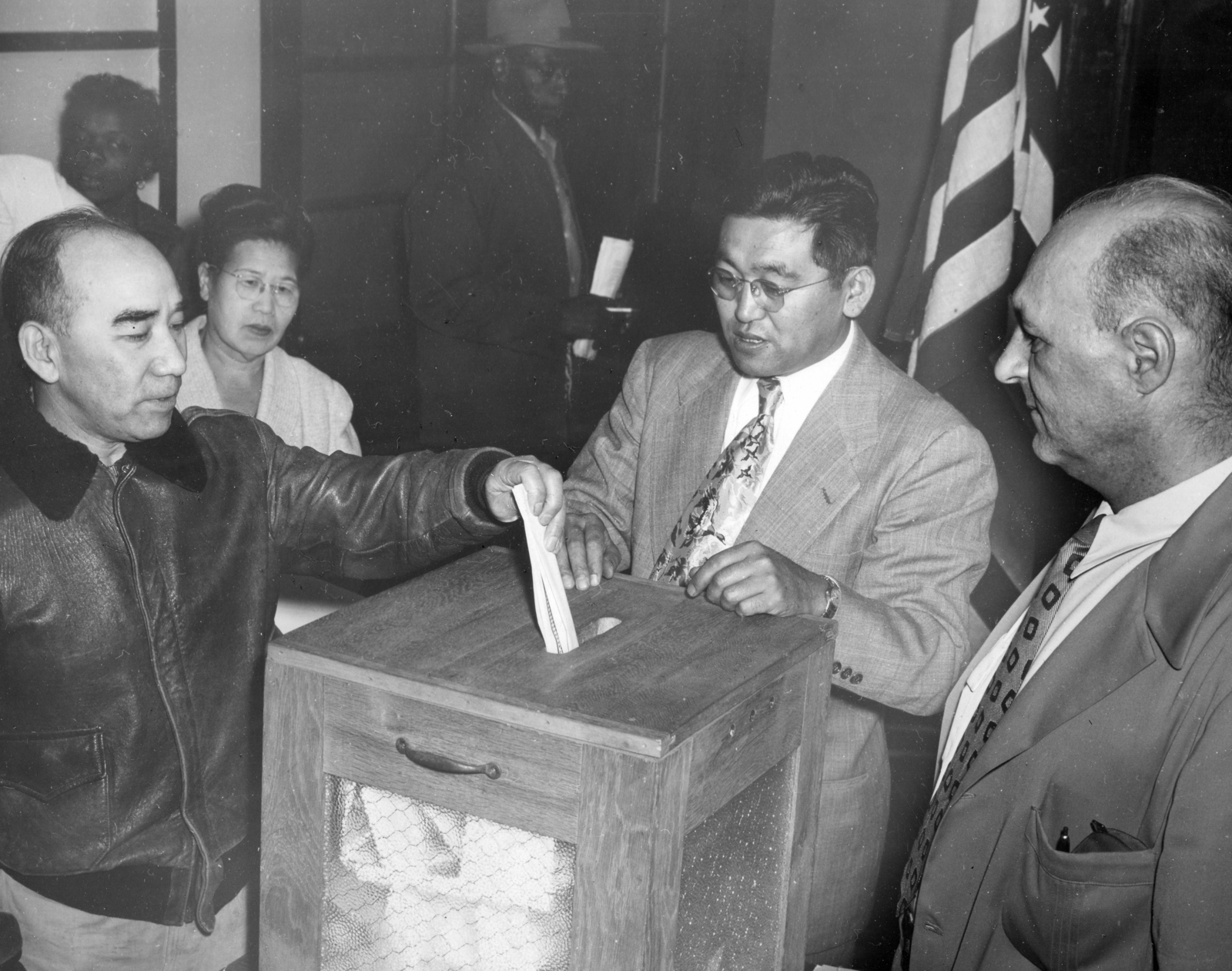 Mitsuzo Fun inserting his paper ballot into the ballot box and being observed by voting officials and other voters